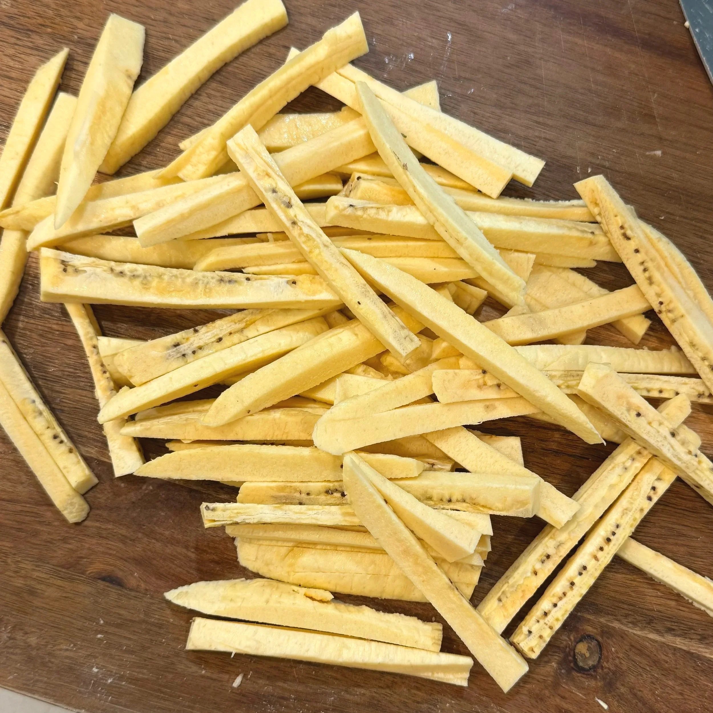 Green plantains peeled and cut into fry-shaped batons on a cutting board, prepped for double-frying.