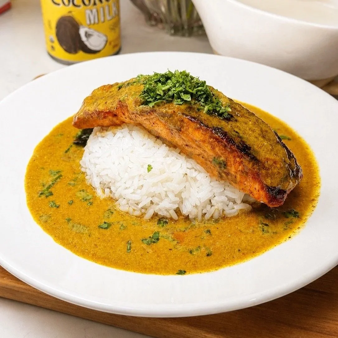  A plate of coconut curry salmon over rice, the seared fillet leaning against the rice with glossy curry sauce pooled around it, finished with fresh cilantro and a lime wedge. 