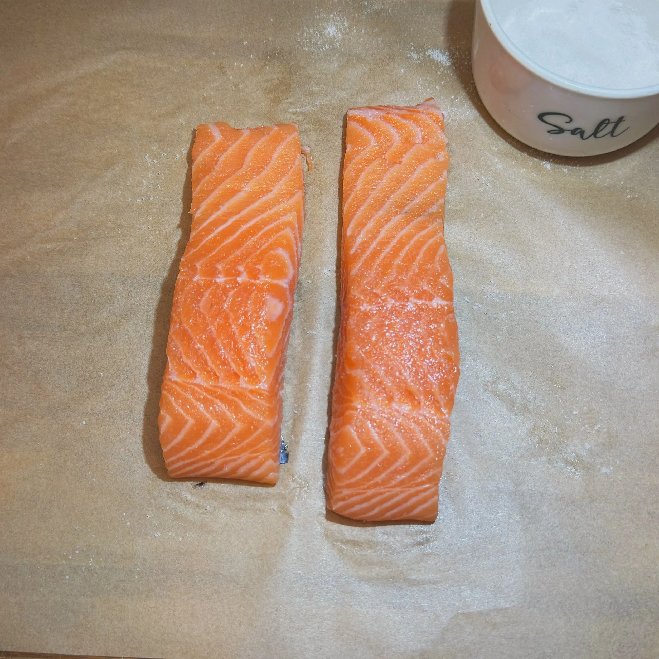  Two skin-on salmon fillets resting on parchment paper, patted dry and ready to be seasoned before searing. 